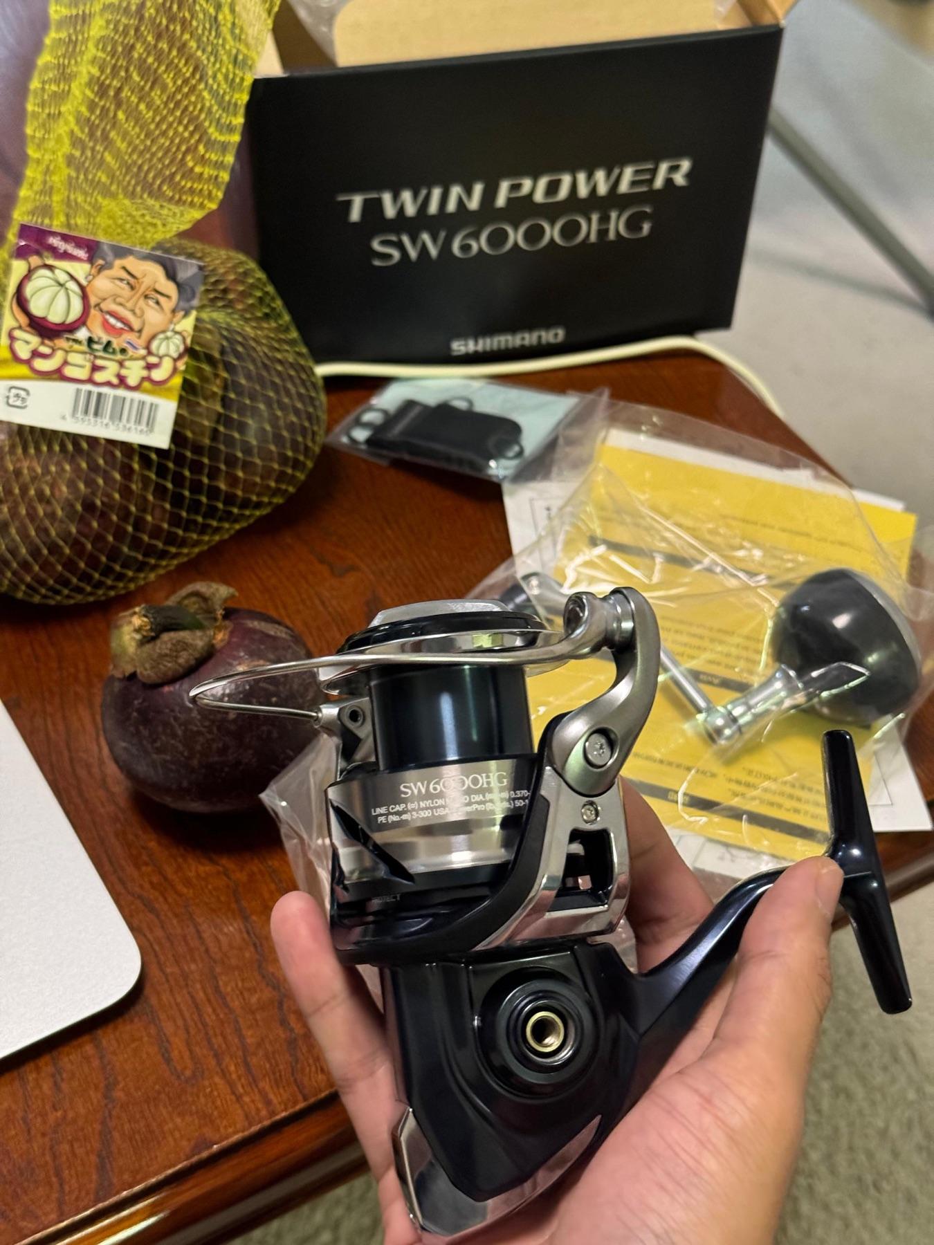 Hand holding a new Shimano Twin Power SW 6000HG spinning reel beside its box and a bag of mangosteen